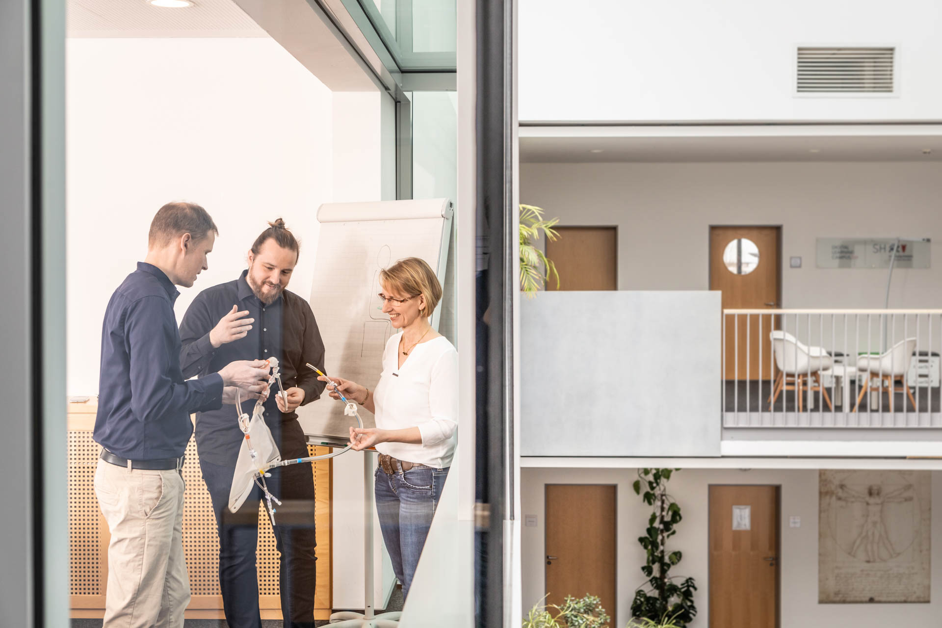 Drei Personen stehen in einem modernen Büro vor einem Flipchart und arbeiten gemeinsam an einem Modell aus Papier und Schnüren, sichtbar durch eine Glaswand im hellen Atrium.